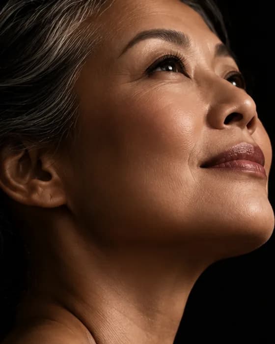 A woman in her fifties with silver-streaked hair, lit against a dark background and looking upward.